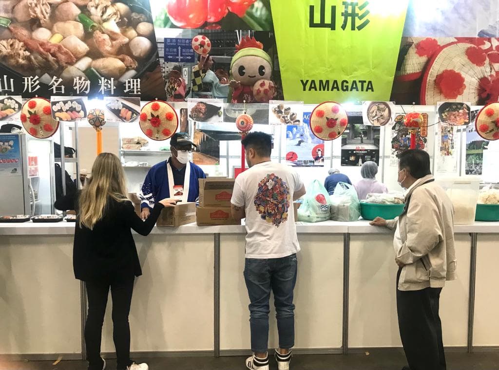 Voluntário atendendo visitantes no balcão do estande da Yamagata em 2022
