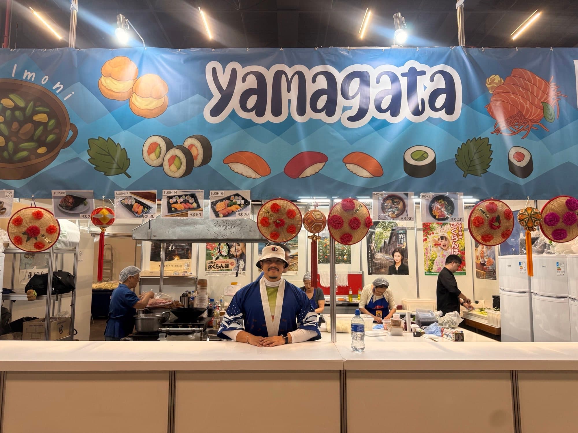 Voluntário em happi coat atendendo no balcão do estande da Yamagata em 2025
