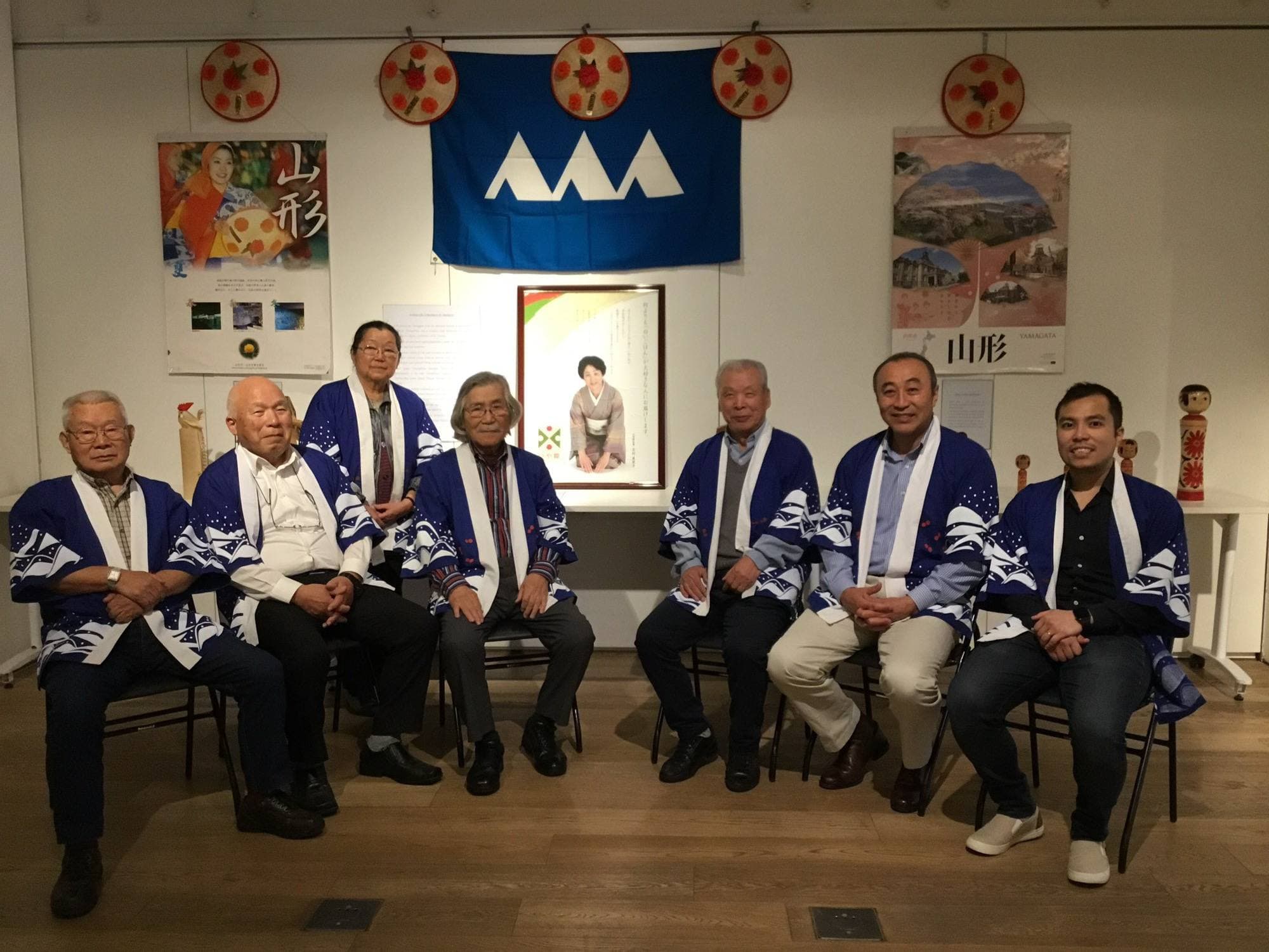 Diretoria da associação em happi coats na divulgação de Yamagata na Japan House, em 2019, com a bandeira da província ao fundo